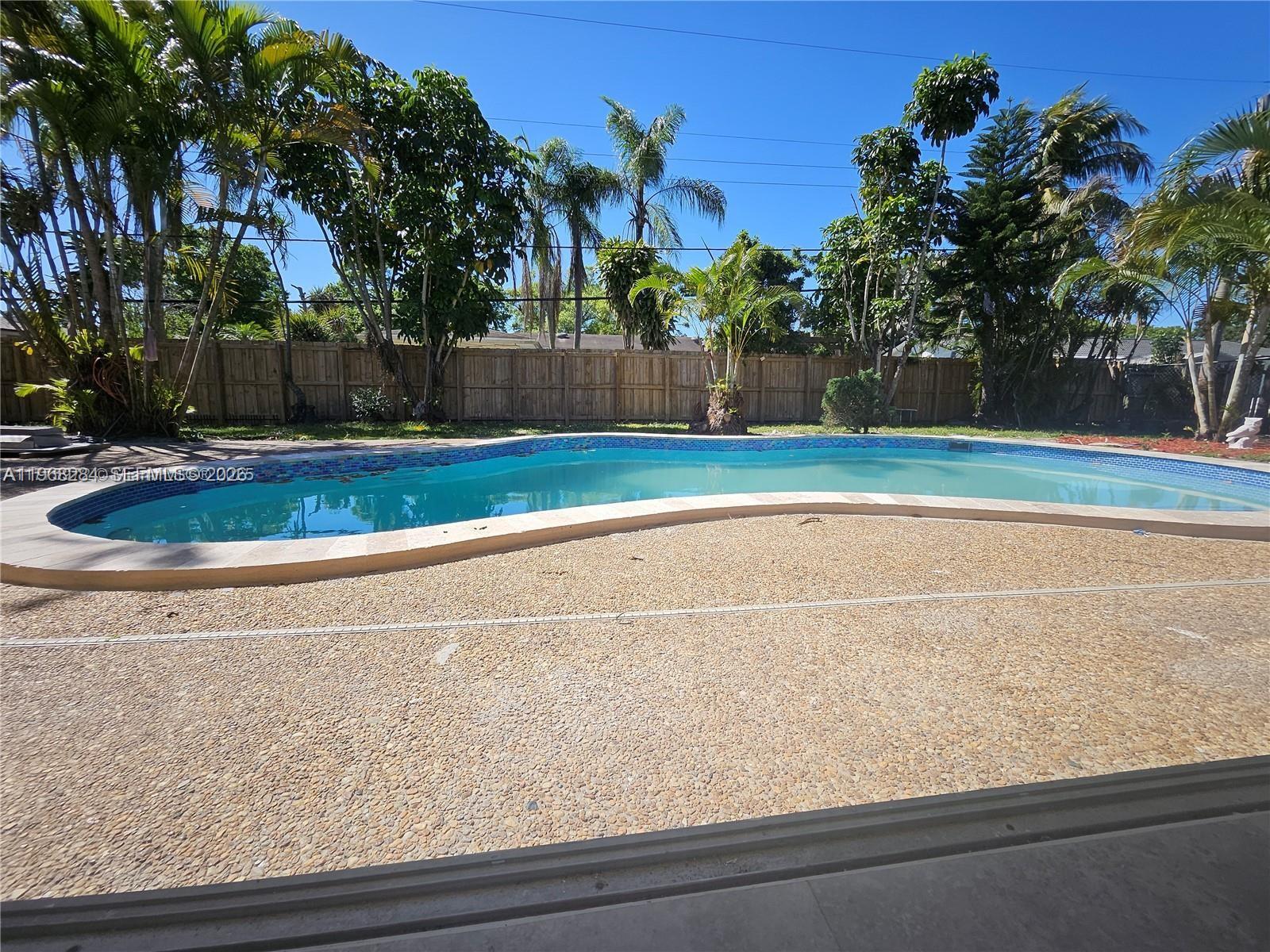 A backyard features a curved swimming pool surrounded by a textured concrete deck and trees — 221 SW 53rd Ave, Plantation FL 33317, PLANTATION, FL