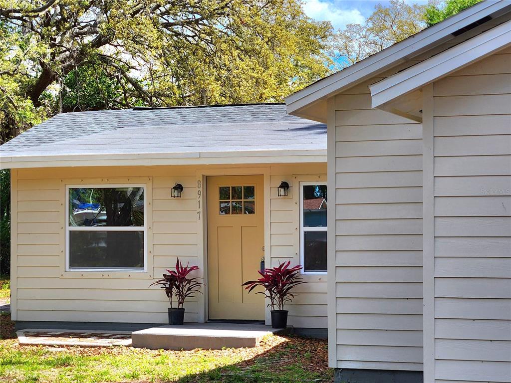 A light-colored house exterior features a yellow front door, two windows, and two potted plants — 8917 N HIGHLAND AVENUE, TAMPA, FL