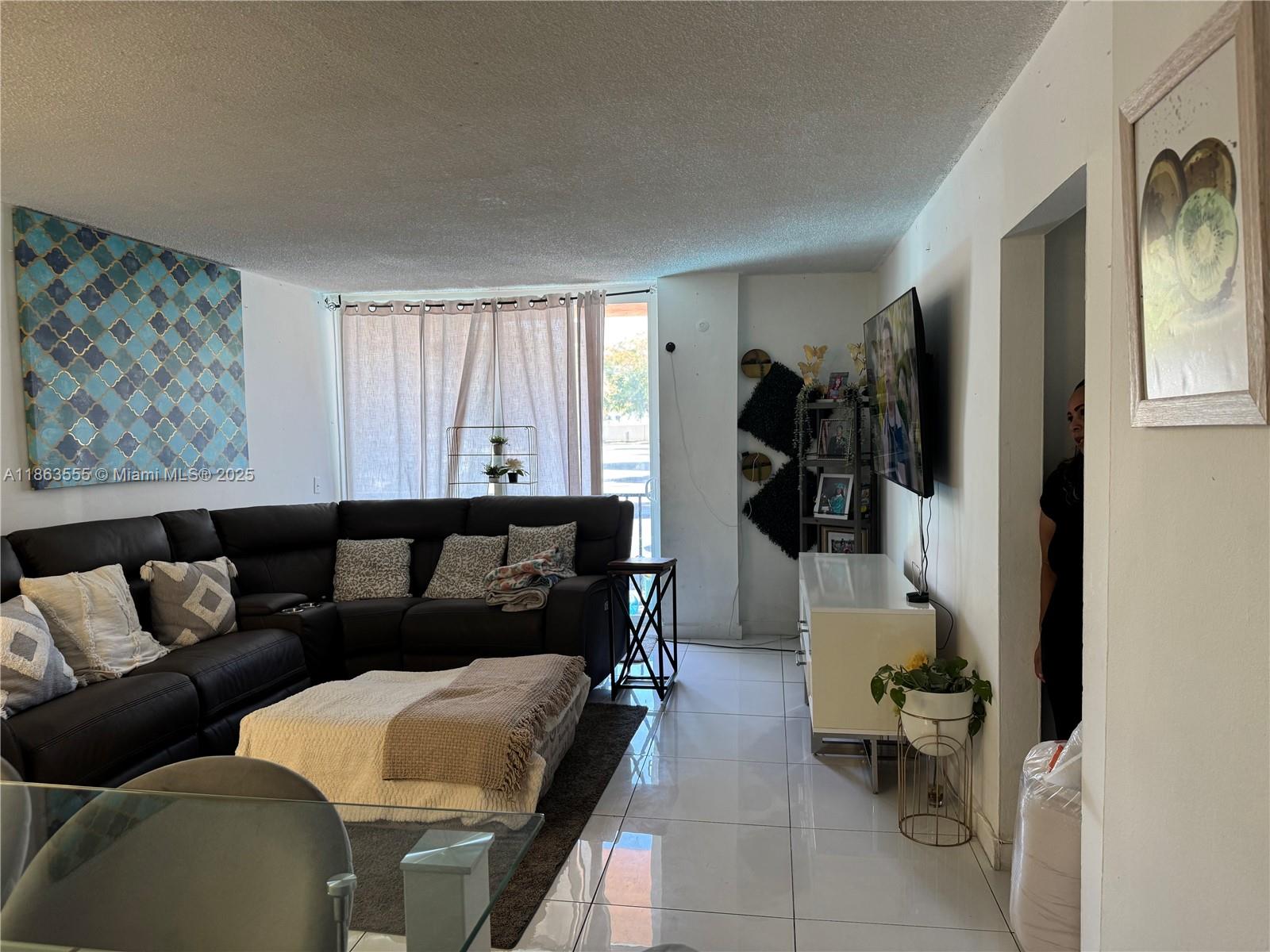 A living room features a dark sectional sofa, white tile floors, and a wall-mounted television — 2075 NE 164th St # 105, North Miami Beach FL 33162, NORTH MIAMI BEACH, FL