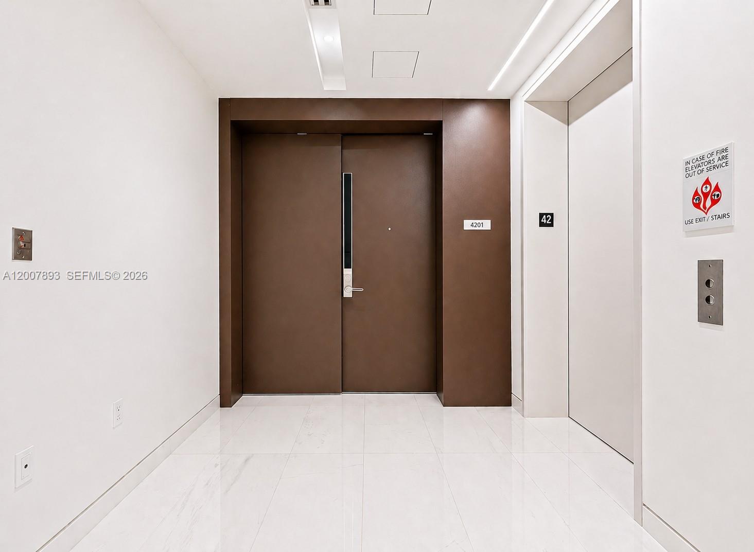 A modern hallway features a dark brown double door, white marble floors, and elevator controls — 300 Biscayne Boulevard Way # 4201E, Miami FL 33131, MIAMI, FL