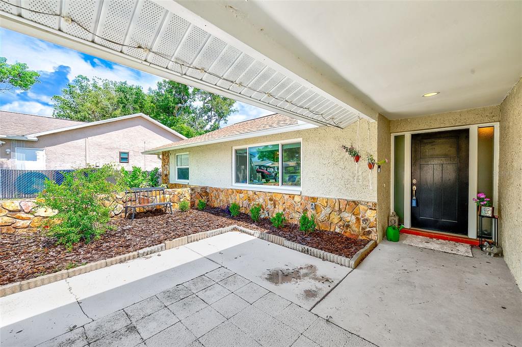 A covered concrete patio features a stone-accented wall, a bench, and a black front door — 8741 HONEYCOMB DRIVE, PORT RICHEY, FL