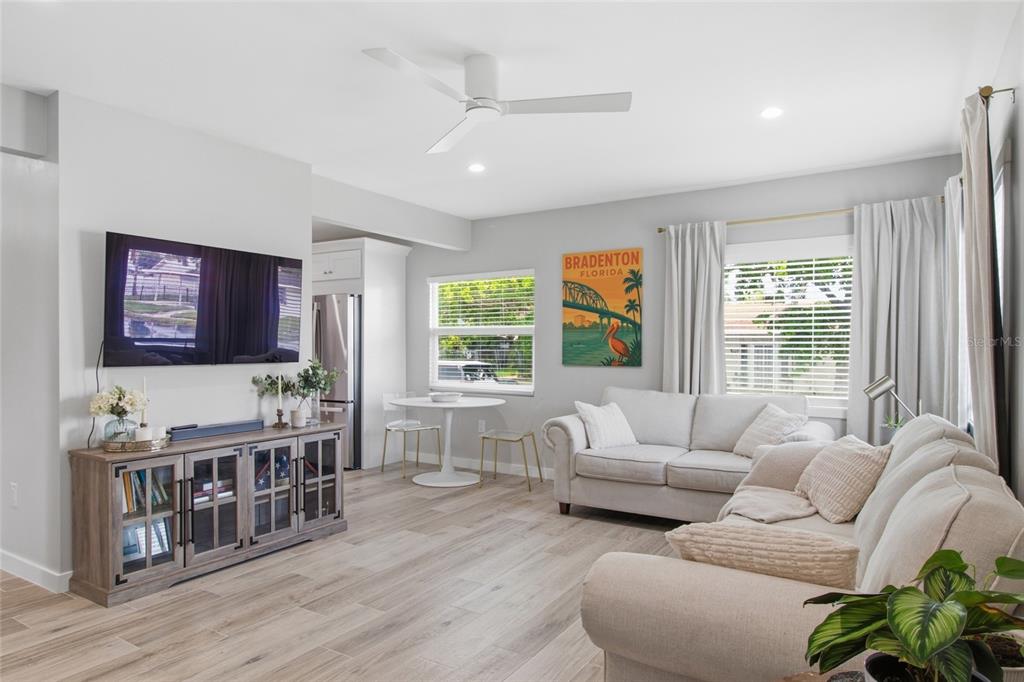 A bright living room features light wood-look flooring, a white sofa, and a television console — 1902 SAINT GEORGE DRIVE, BRADENTON, FL