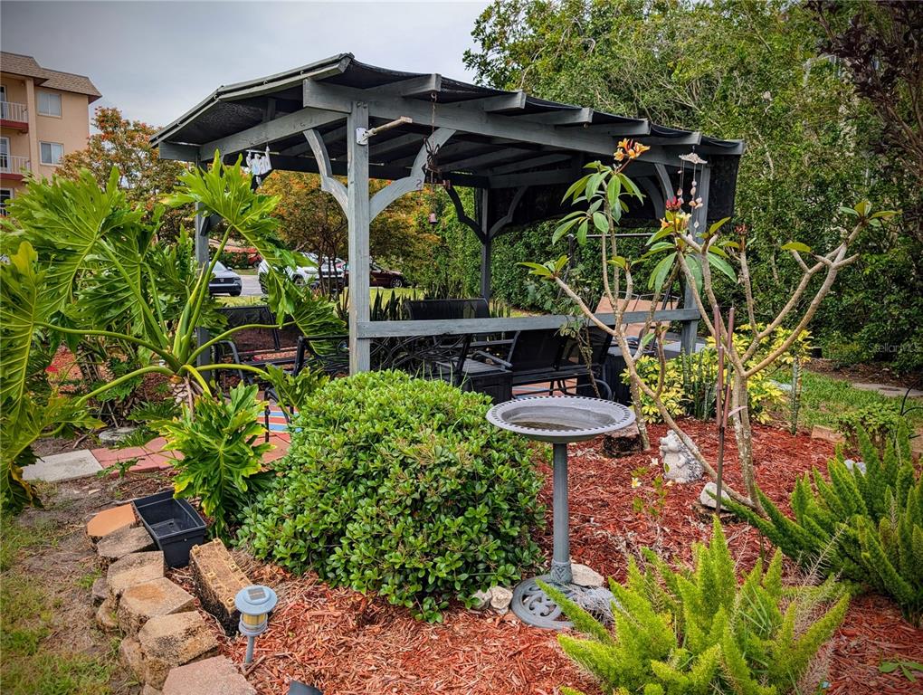 A wooden gazebo sits in a landscaped garden with a birdbath and various plants — 4127 HERON WAY UNIT E-111, BRADENTON, FL