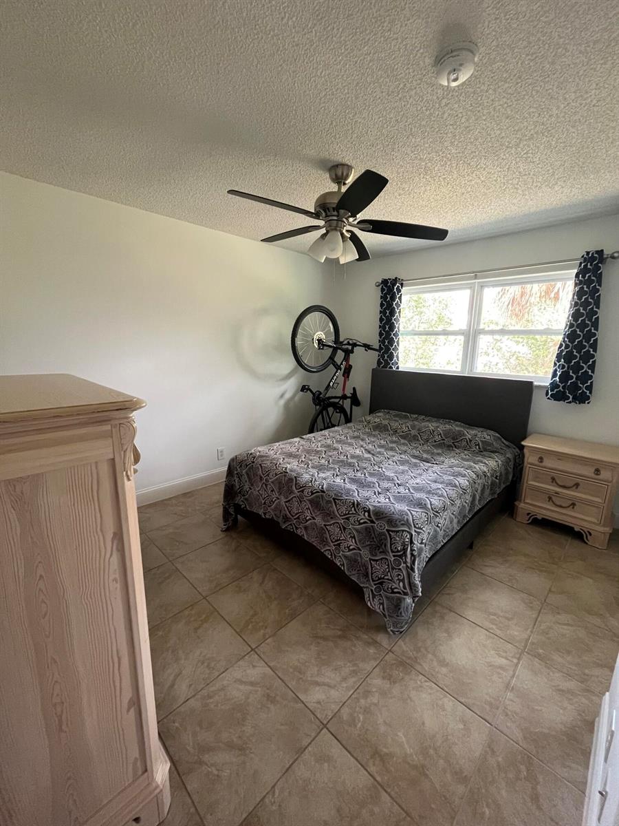 A bedroom features tile flooring, a ceiling fan, a bed, and a bicycle stored inside — 1979 Juno Road, North Palm Beach FL 33408, NORTH PALM BEACH, FL