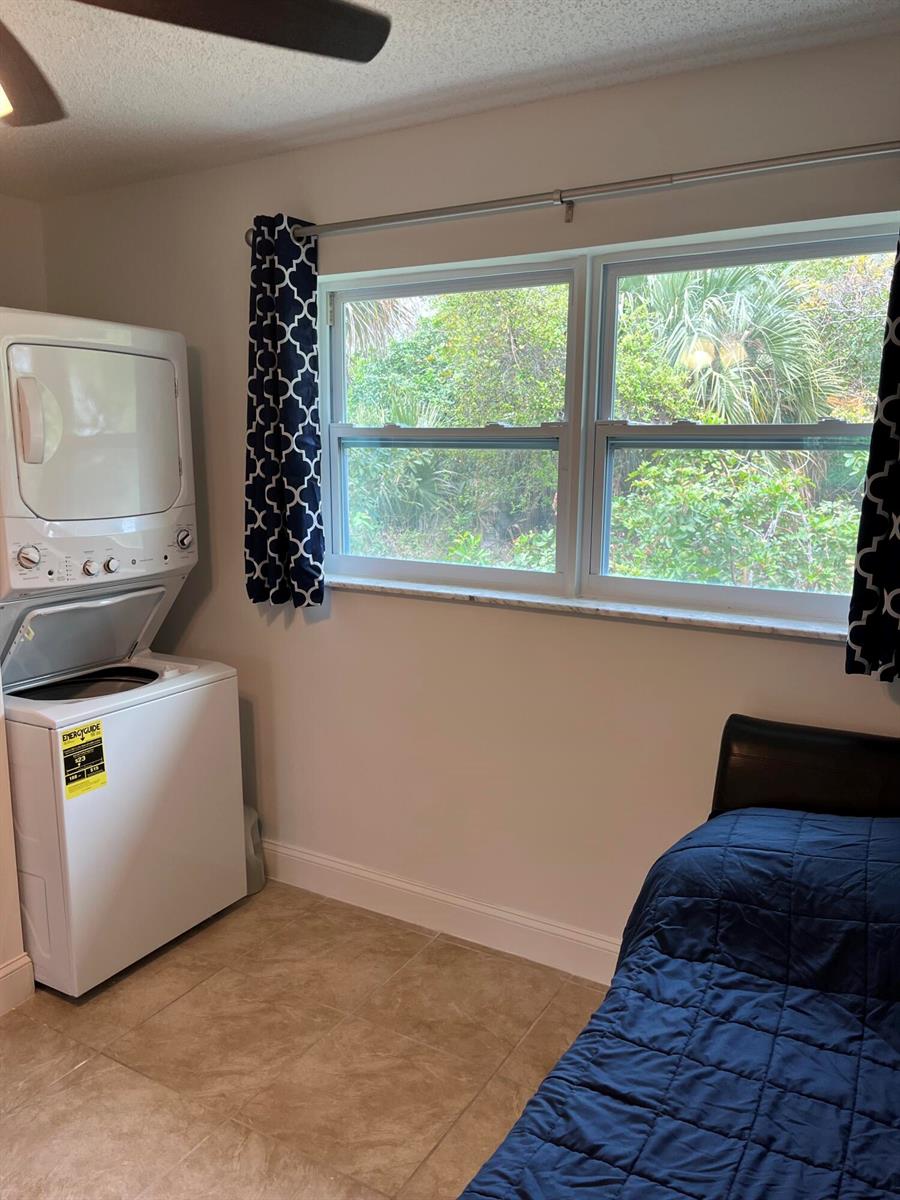 A bedroom with tile flooring features a stacked washer and dryer unit and a window — 1979 Juno Road, North Palm Beach FL 33408, NORTH PALM BEACH, FL