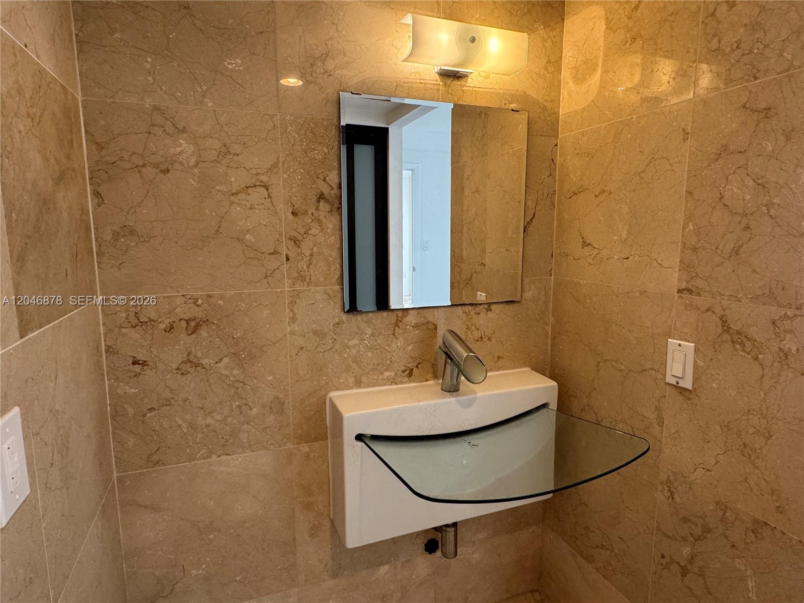 A small bathroom features beige marble wall tiles, a wall-mounted sink, and a glass shelf — 1345 West Ave # 202, Miami Beach FL 33139, MIAMI BEACH, FL