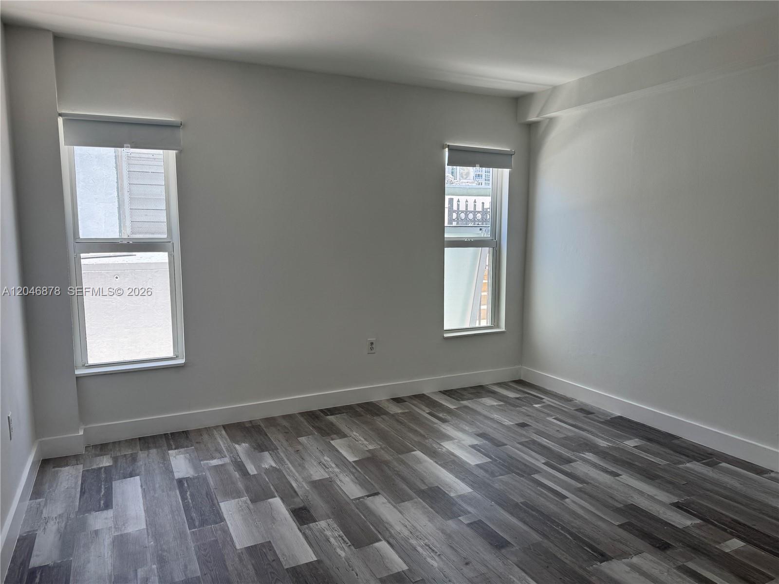 An empty room features white walls, gray wood-look flooring, and two windows with blinds — 1345 West Ave # 202, Miami Beach FL 33139, MIAMI BEACH, FL