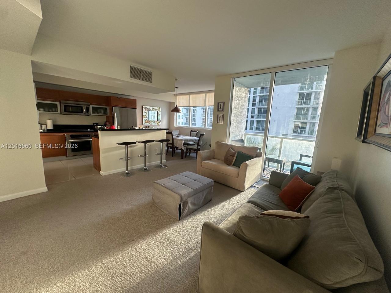 A living room with carpeted floors, a sofa, an ottoman, and an open kitchen area — 253 NE 2nd St # 1402, Miami FL 33132, MIAMI, FL