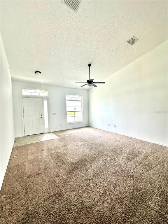 A spacious, empty living room features beige carpeting, white walls, and a ceiling fan — 102 AURELIA COURT, KISSIMMEE, FL