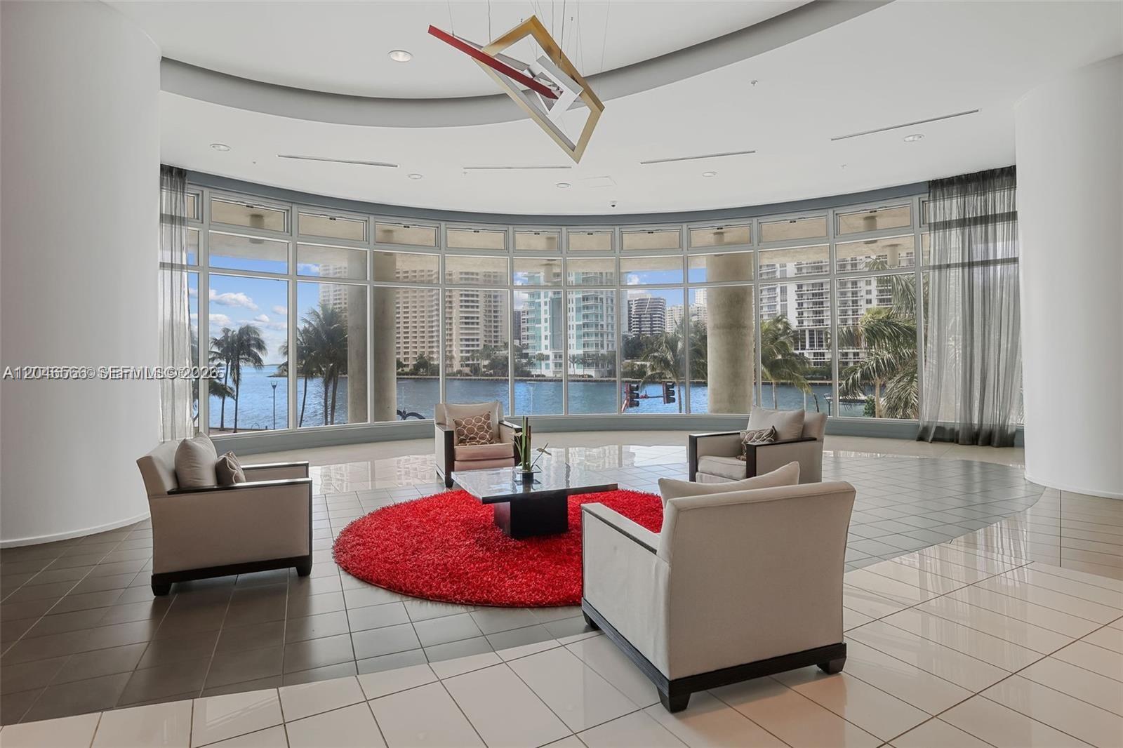A circular seating area with a red rug overlooks a waterfront city view through windows — 300 S Biscayne Blvd # 2603, Miami FL 33131, MIAMI, FL