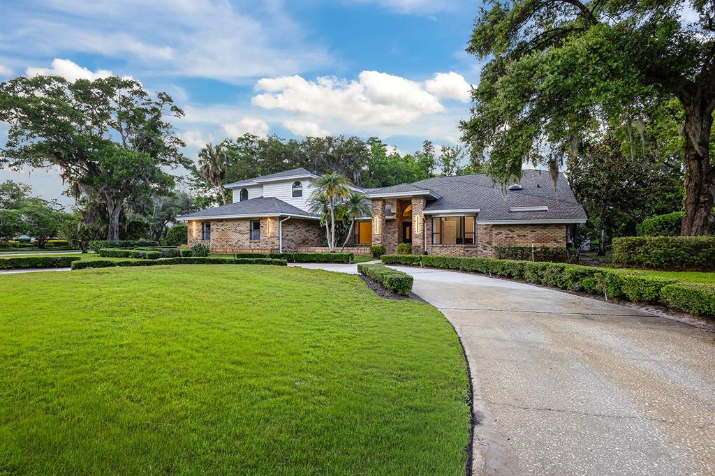 A brick house with a curved driveway, manicured hedges, and large trees under a blue sky — 2901 ST ANDREWS BOULEVARD, TARPON SPRINGS, FL