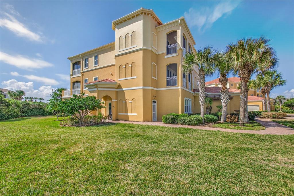 A three-story yellow stucco building with arched windows and balconies stands on a grassy lawn — 160 AVENUE DE LA MER UNIT 1903, PALM COAST, FL