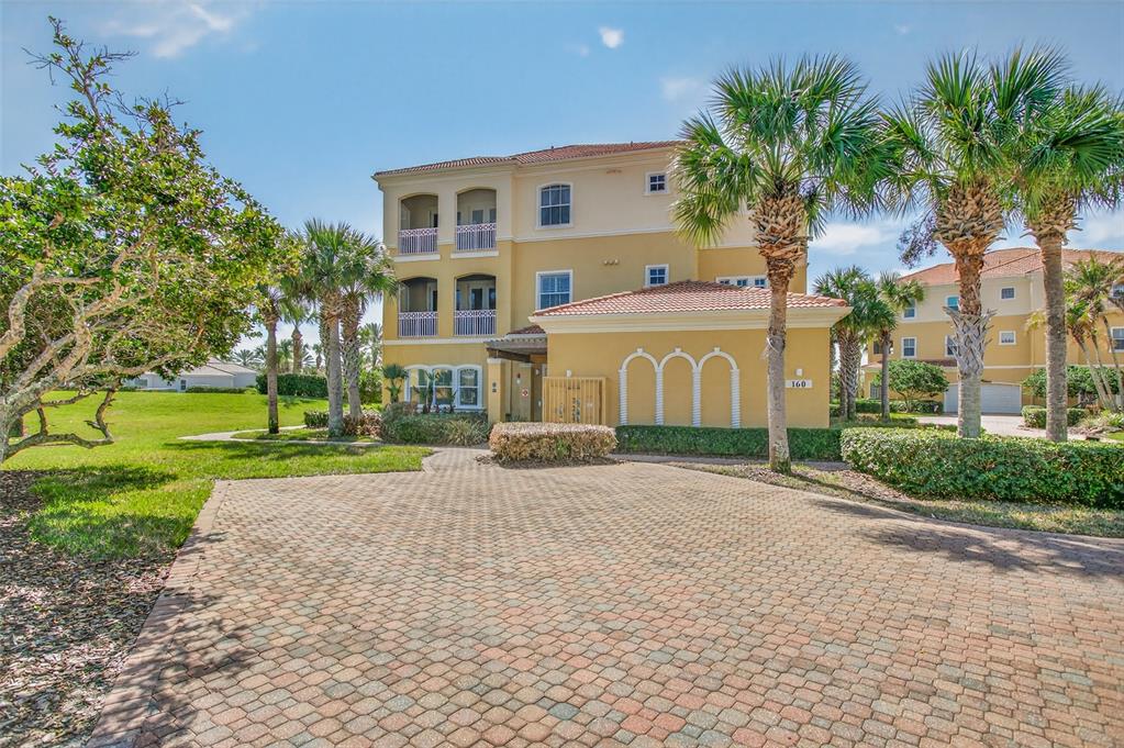A three-story yellow stucco building with a paved driveway and palm trees in front — 160 AVENUE DE LA MER UNIT 1903, PALM COAST, FL