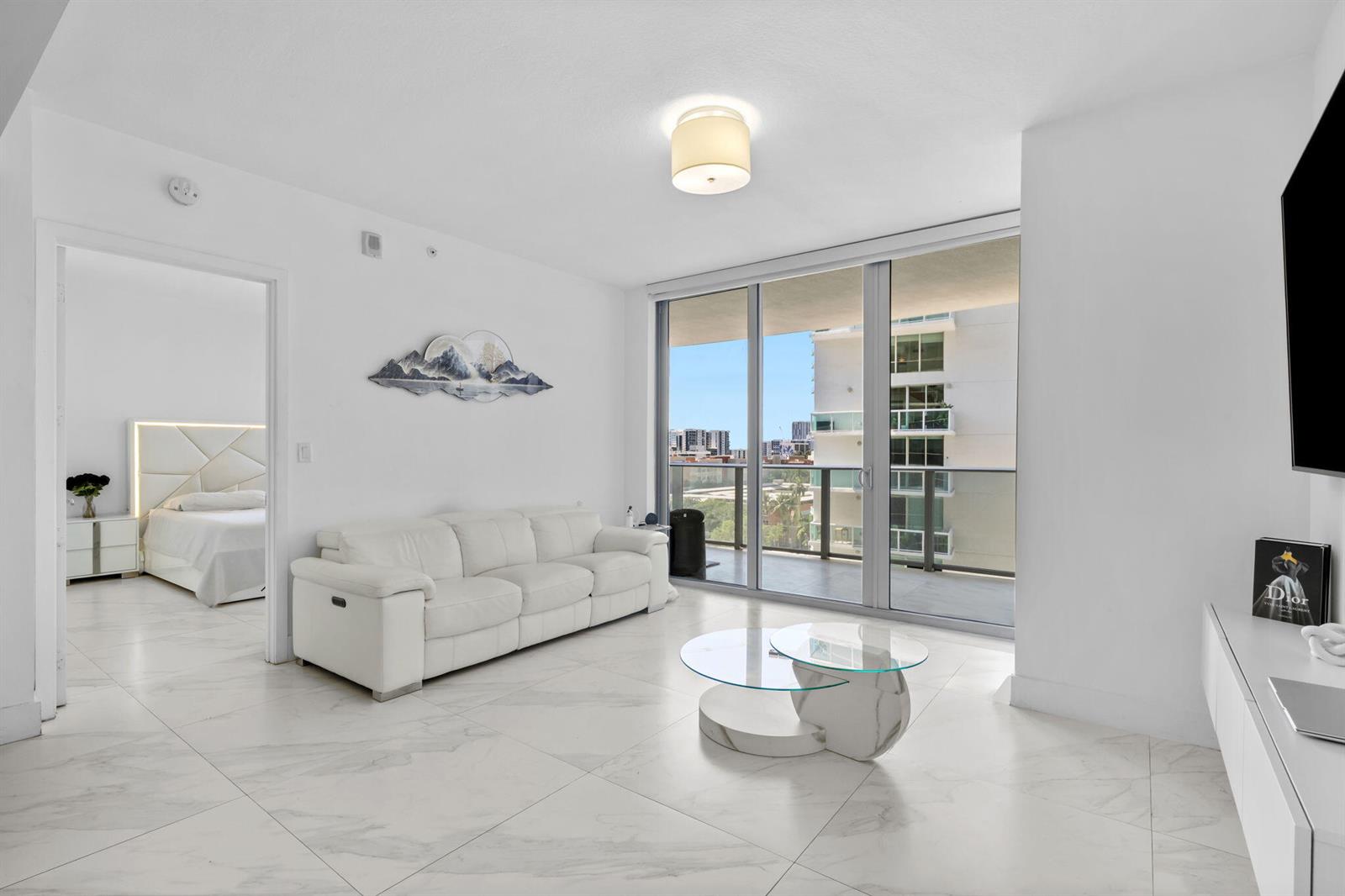 A bright living room with white tile floors, a white sofa, and a glass coffee table — 488 NE 18th Street # 905, Miami FL 33132, MIAMI, FL