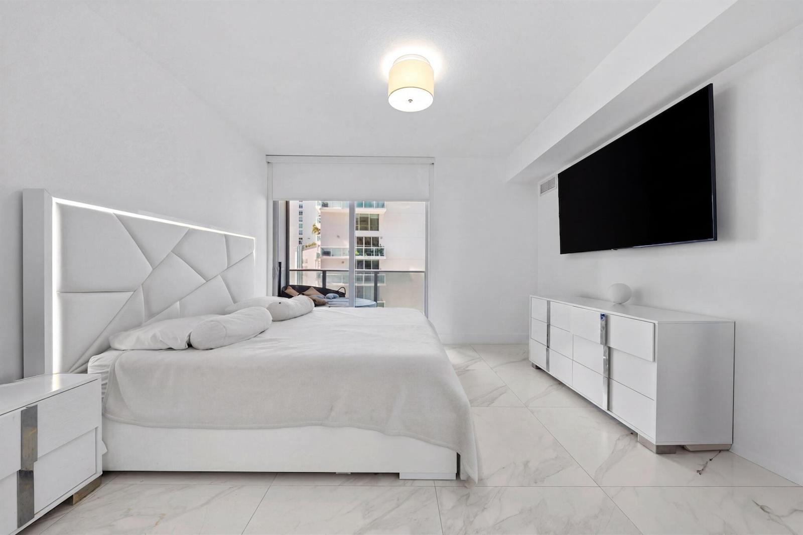 A modern white bedroom features a tufted headboard, marble-look tile floors, and a wall-mounted television — 488 NE 18th Street # 905, Miami FL 33132, MIAMI, FL