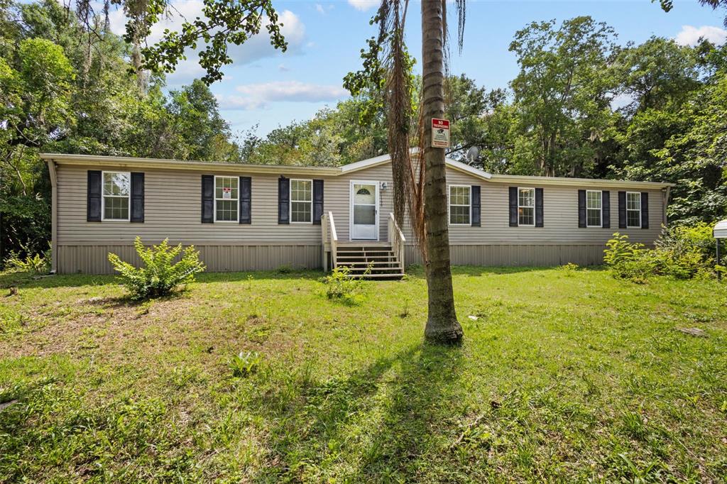 A single-story manufactured home with beige siding sits on a grassy lot surrounded by trees — 21390 NW 59TH AVENUE, MICANOPY, FL