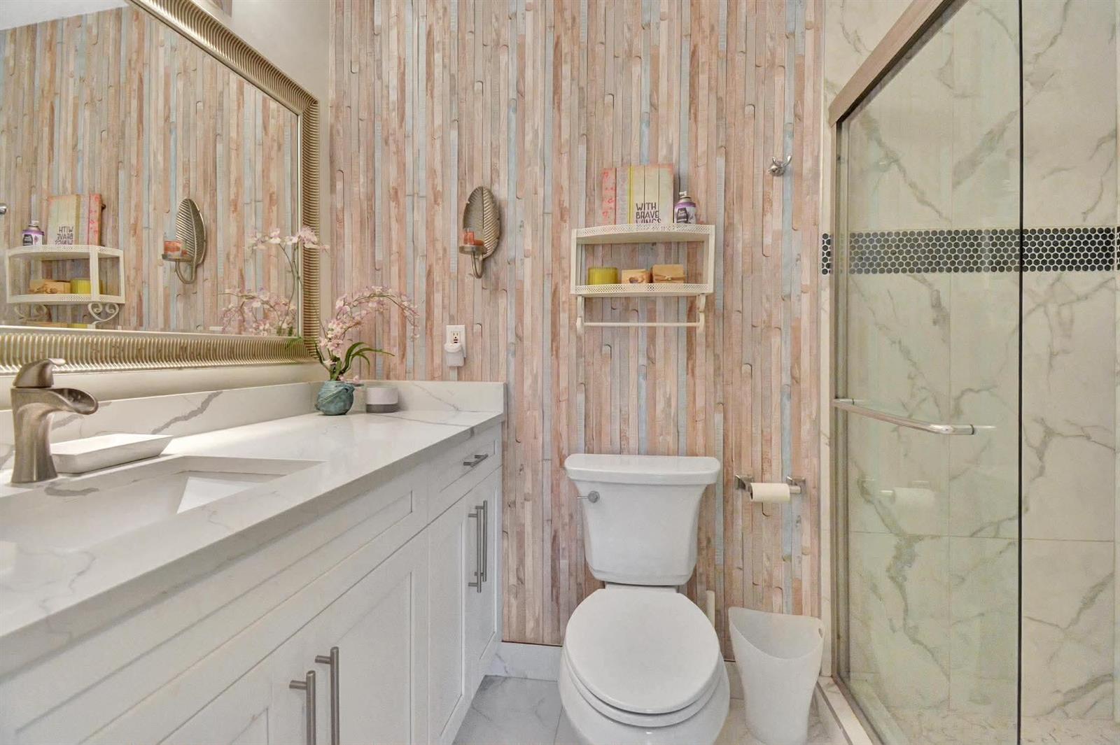 A bathroom features a white vanity, a toilet, a glass shower, and wood-look vertical wallpaper — 1232 Club Drive W # D, Delray Beach FL 33445, DELRAY BEACH, FL