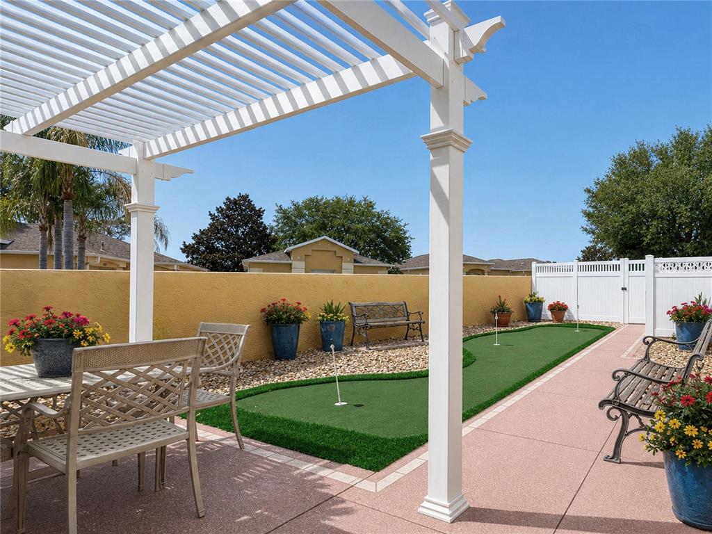 A backyard patio features a white pergola, a putting green, and potted flowers — 853 WINCHESTER COURT, THE VILLAGES, FL