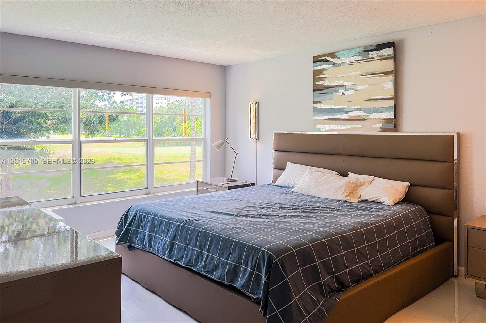 A bedroom features a large brown upholstered bed, a window with park views, and artwork — 3200 N Palm Aire Dr # 207, Pompano Beach FL 33069, POMPANO BEACH, FL