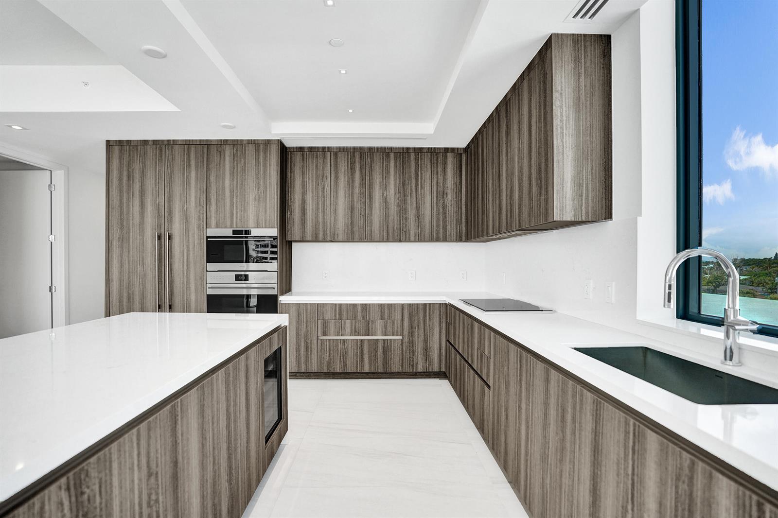A modern kitchen features wood-grain cabinets, white countertops, a central island, and a large window — 305 Briny Avenue # 306, Pompano Beach FL 33062, POMPANO BEACH, FL