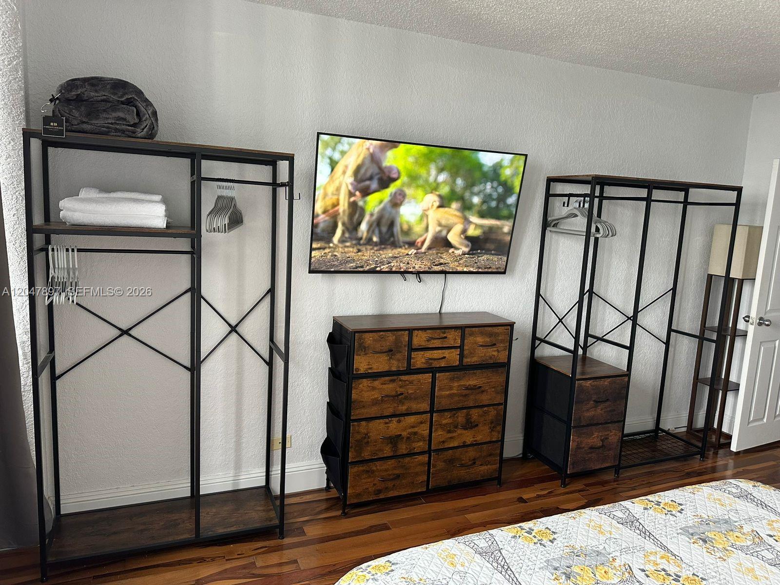 A bedroom features a wall-mounted television, a wooden dresser, and two open metal clothing racks — 1500 Bay Rd # 1408S, Miami Beach FL 33139, MIAMI BEACH, FL