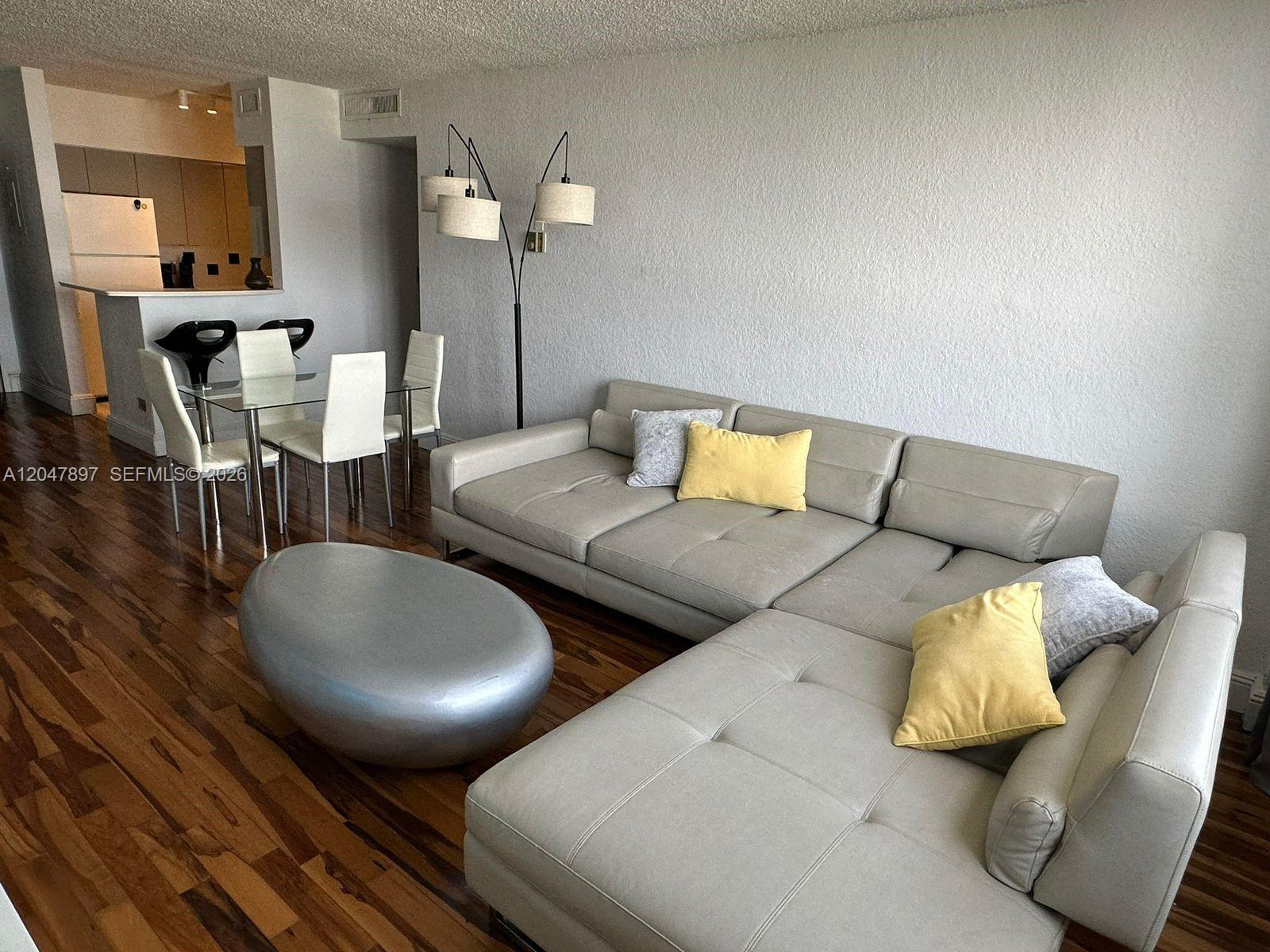 A living room features a light gray sectional sofa, a silver oval ottoman, and dining area — 1500 Bay Rd # 1408S, Miami Beach FL 33139, MIAMI BEACH, FL