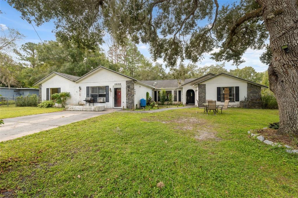 A single-story white house with stone accents, a driveway, and a large tree in front — 18160 SPANGLER AVENUE, BROOKSVILLE, FL