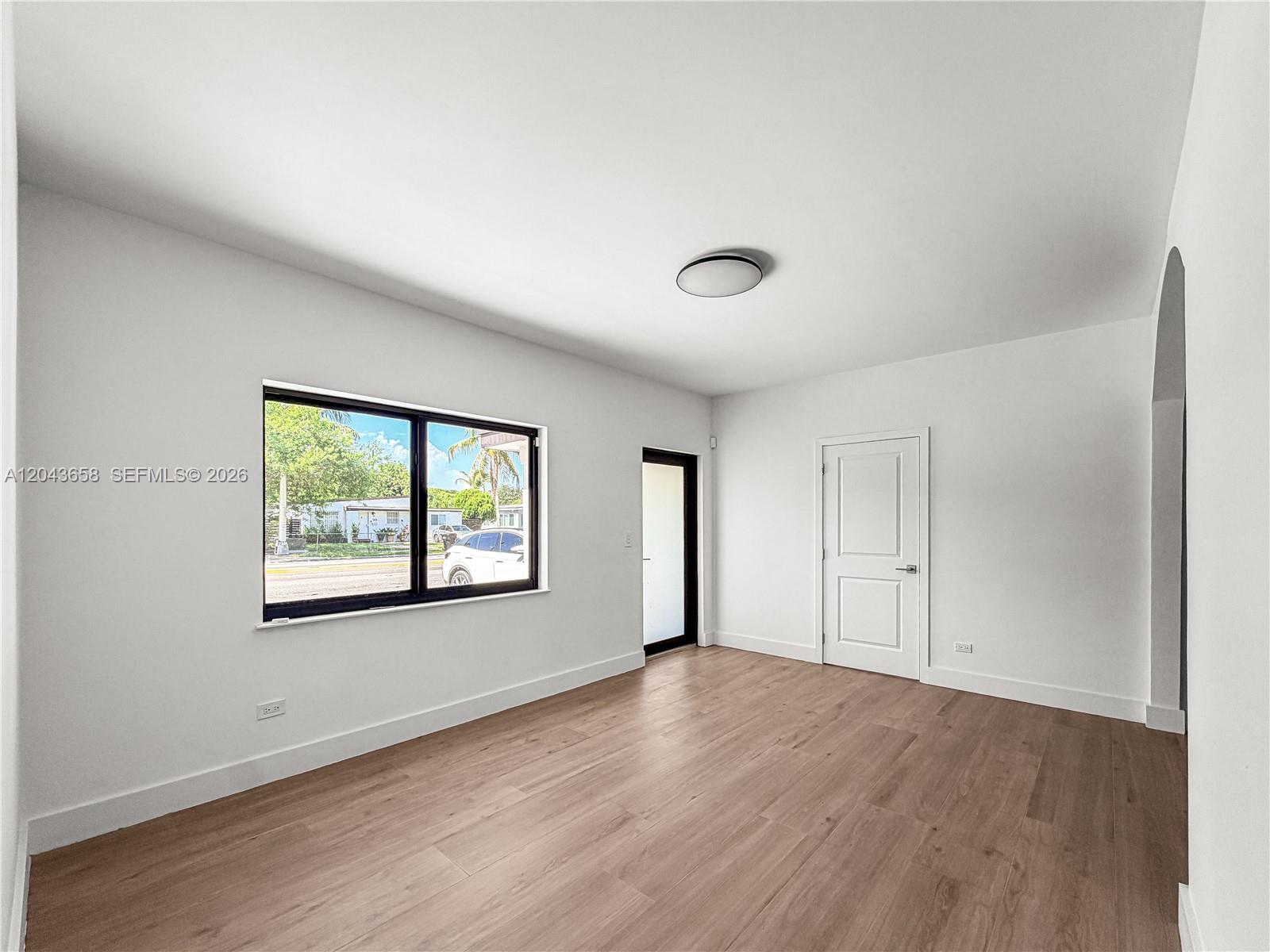 An empty room features white walls, light wood-look flooring, a window, and a door — 55 NW 125th St, North Miami FL 33168, NORTH MIAMI, FL