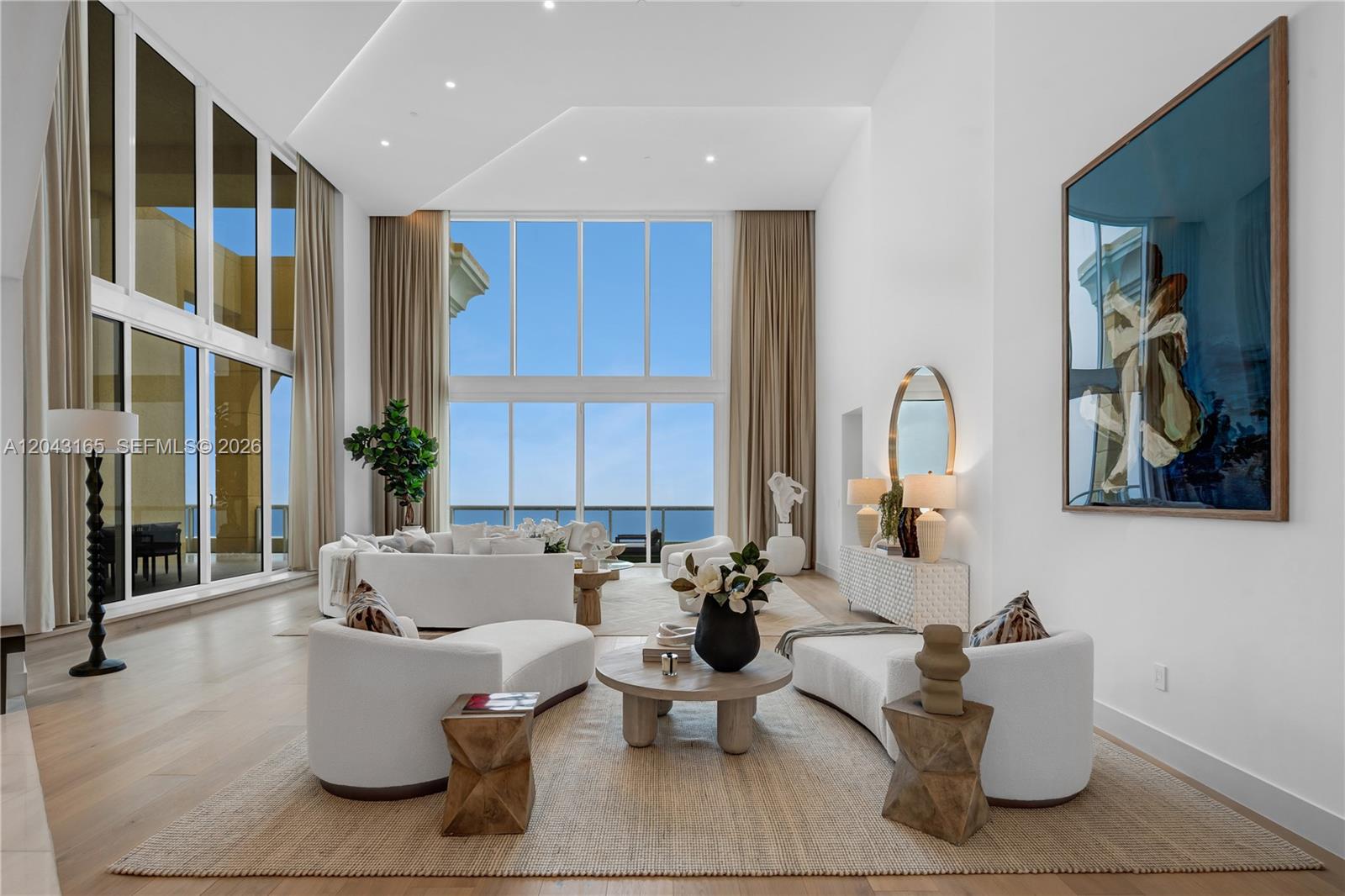 A bright, high-ceilinged living room features white curved furniture, a round wooden table, and floor-to-ceiling windows — 17875 Collins Ave, Sunny Isles Beach FL 33160, SUNNY ISLES BEACH, FL