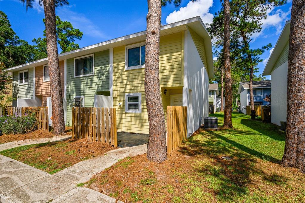 A two-story townhouse with yellow and green siding, a wooden fence, and surrounding pine trees — 2132 SW 39TH DRIVE, GAINESVILLE, FL