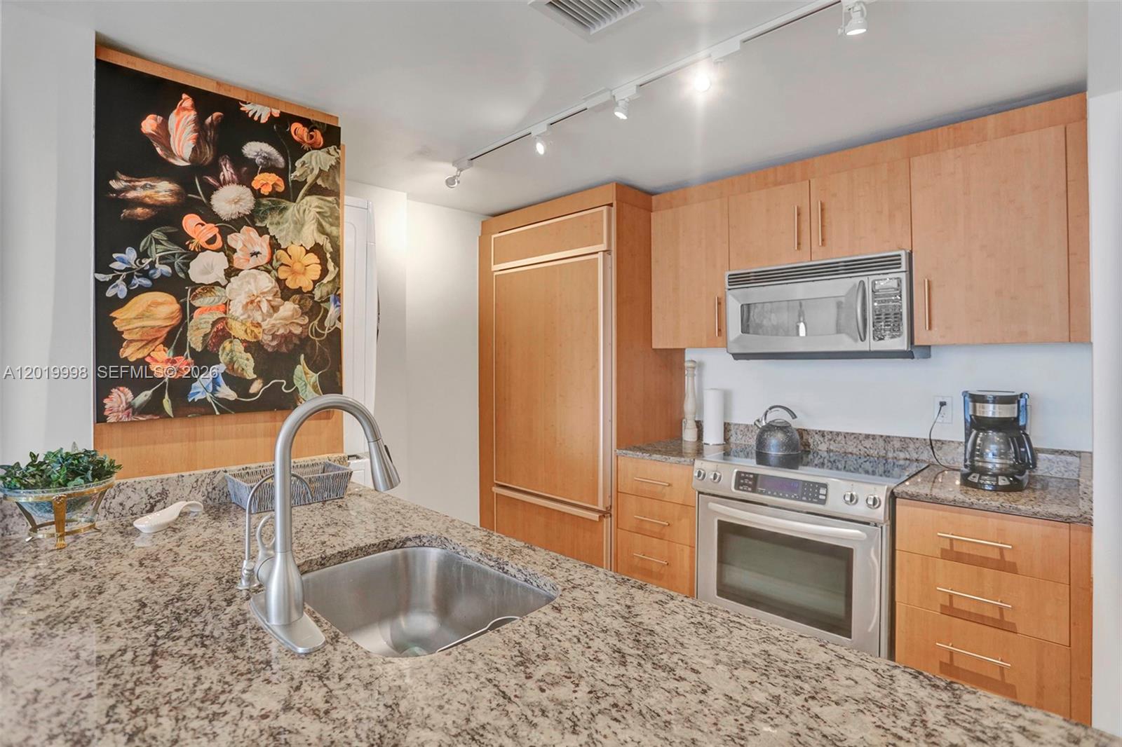 A kitchen features light wood cabinetry, granite countertops, stainless steel appliances, and floral wall art — 10275 Collins Ave # 829, Bal Harbour FL 33154, BAL HARBOUR, FL