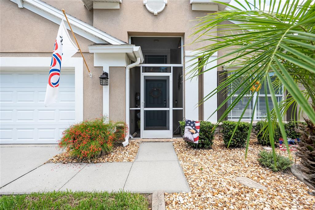 A tan house exterior features a front door with a screen, a garage, and landscaping — 13508 MEADOW GOLF AVENUE, HUDSON, FL