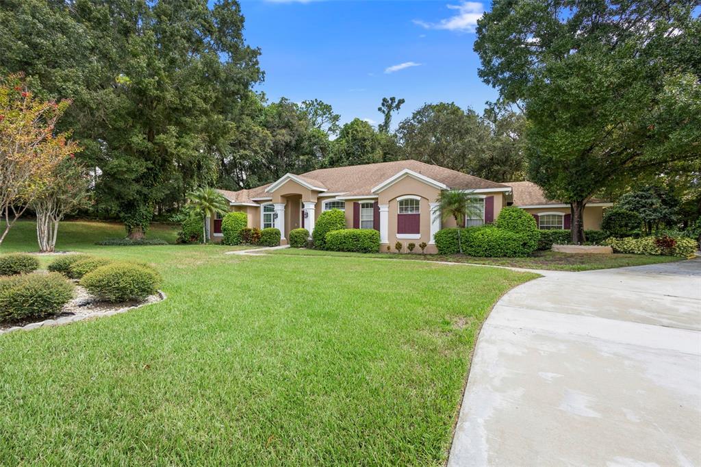 A tan single-story house with a concrete driveway and manicured landscaping sits under large trees — 1621 TIMBER HILLS DRIVE, DELAND, FL