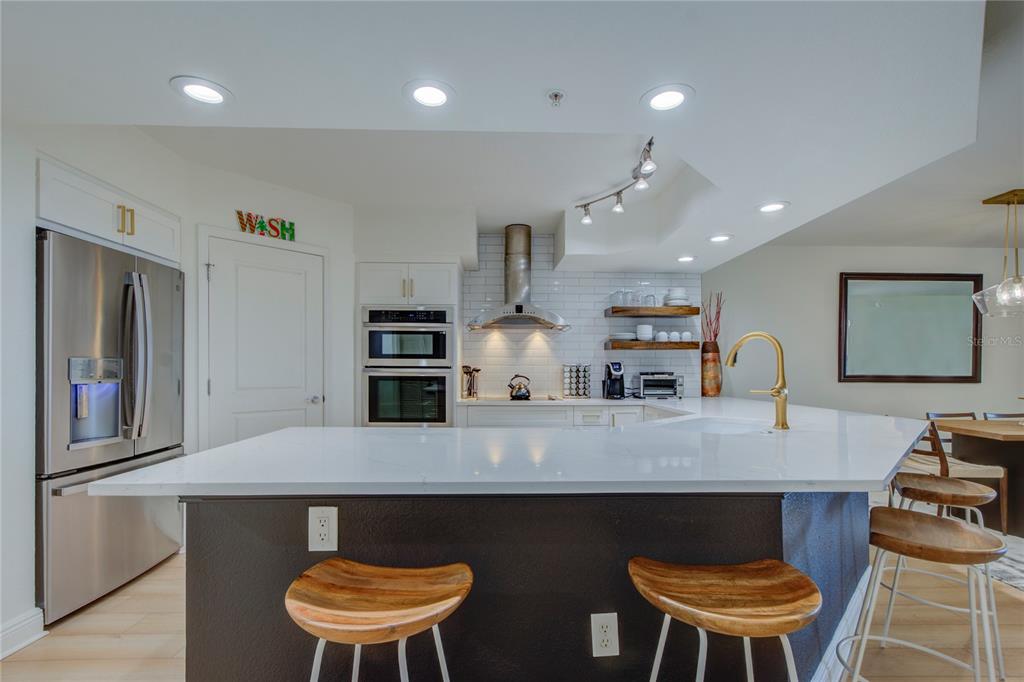 A modern kitchen features a white quartz island, dark cabinetry, stainless steel appliances, and barstools — 5353 GULF BOULEVARD UNIT A502, ST PETE BEACH, FL