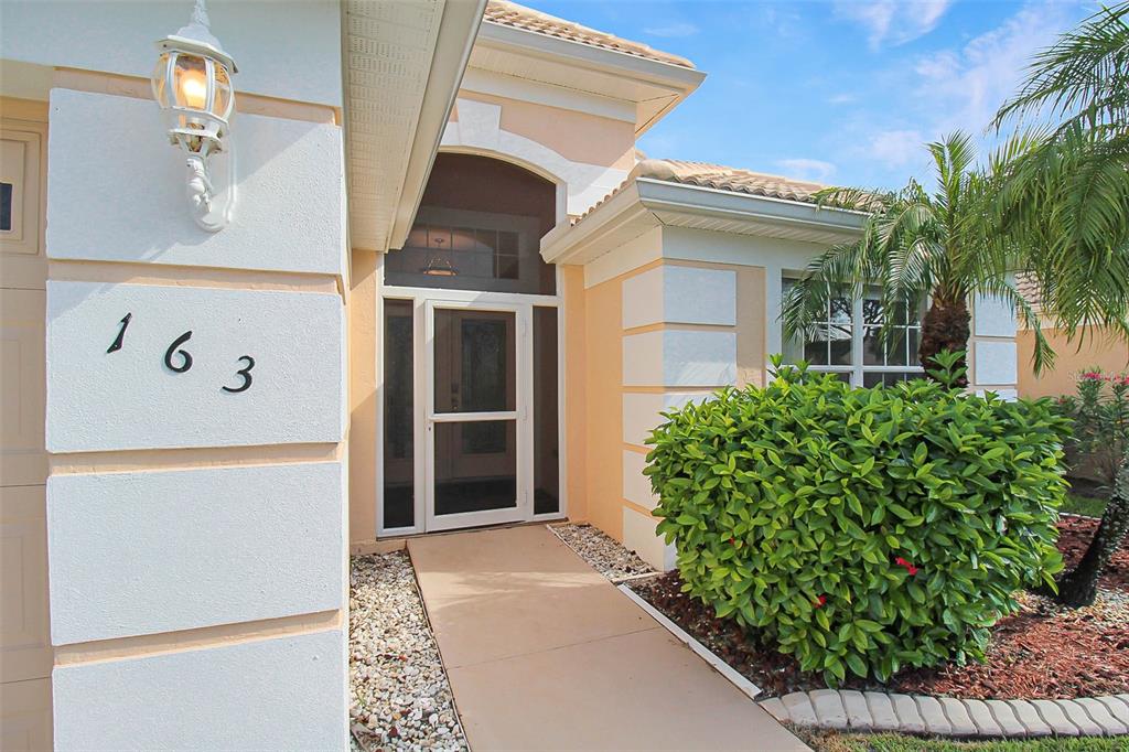 The exterior of a house features a beige stucco facade, a screen door, and house numbers — 163 BRAEMAR AVENUE, VENICE, FL