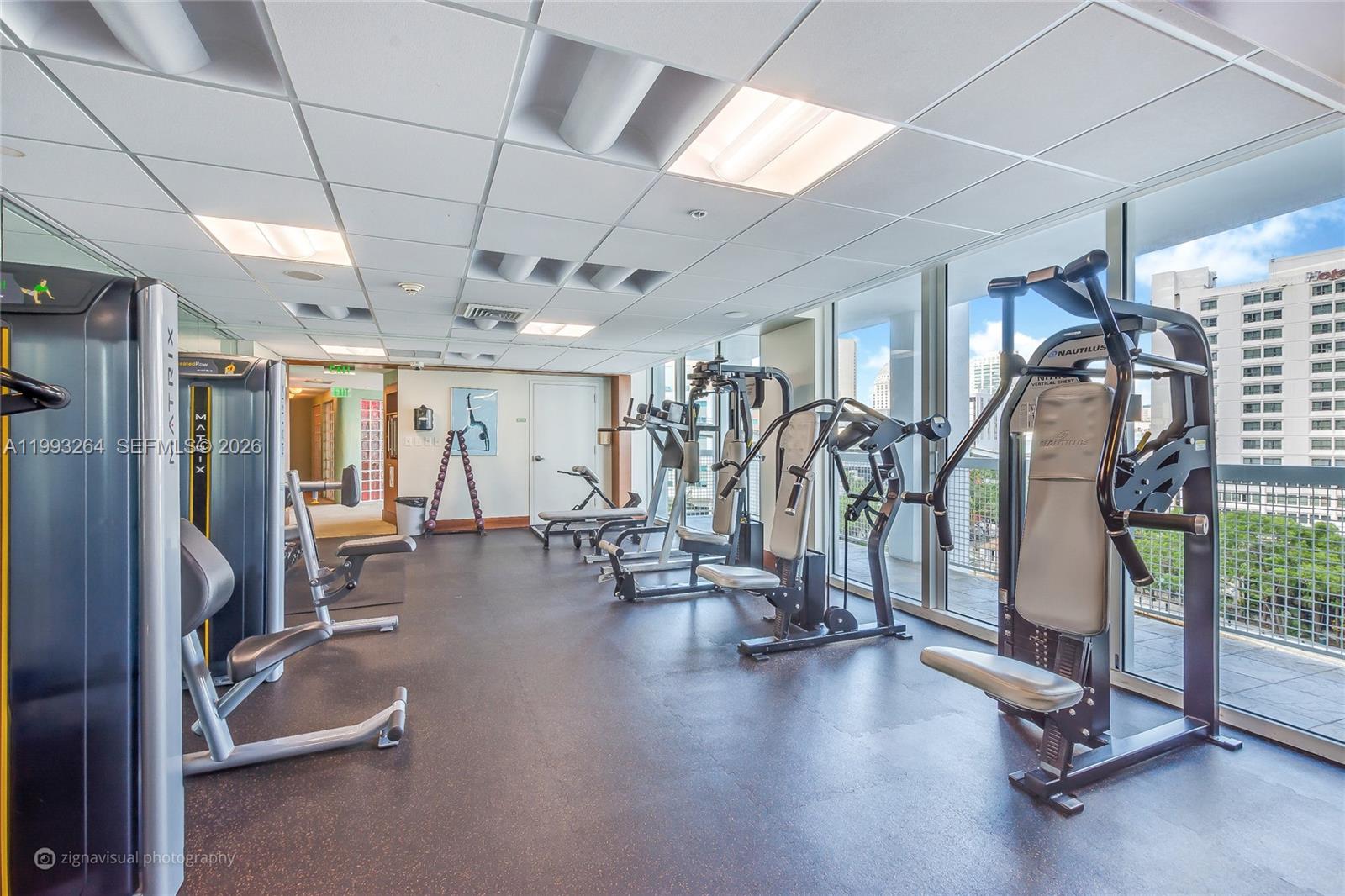 A gym with various exercise machines, rubber flooring, and large windows overlooking a city building — 31 SE 5th St # 2708, Miami FL 33131, MIAMI, FL