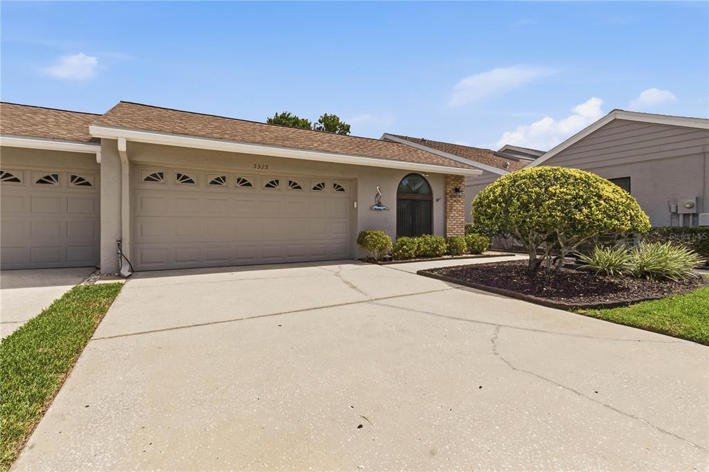 A single-story house exterior features a beige stucco facade, a two-car garage, and landscaping — 5515 SALEM SQUARE DRIVE S, PALM HARBOR, FL