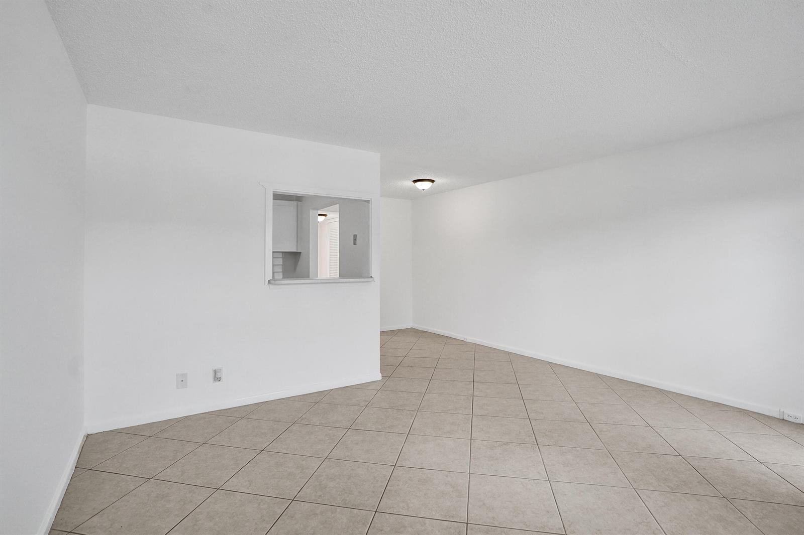 An empty room with white walls, beige tile flooring, and a kitchen pass-through window — 91 Tilford, Deerfield Beach FL 33442, DEERFIELD BEACH, FL