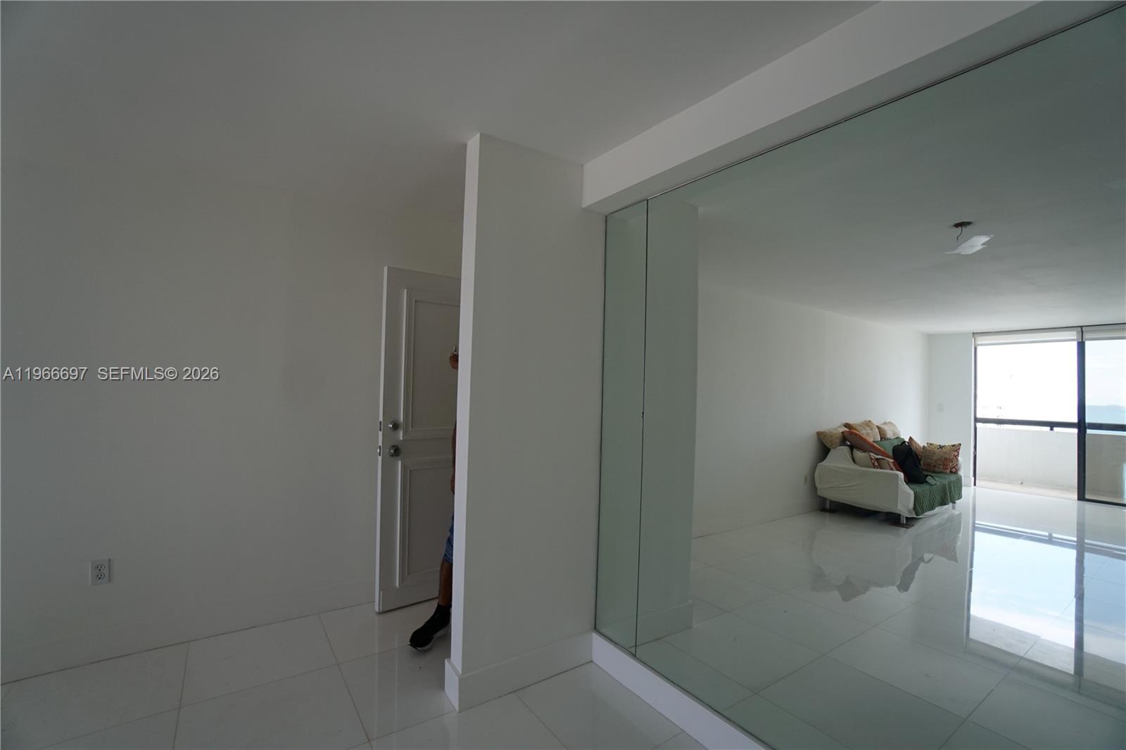 A white-walled room features a glass-walled partition, white tile flooring, and a sofa near windows — 1775 Washington Ave # 11C, Miami Beach FL 33139, MIAMI BEACH, FL
