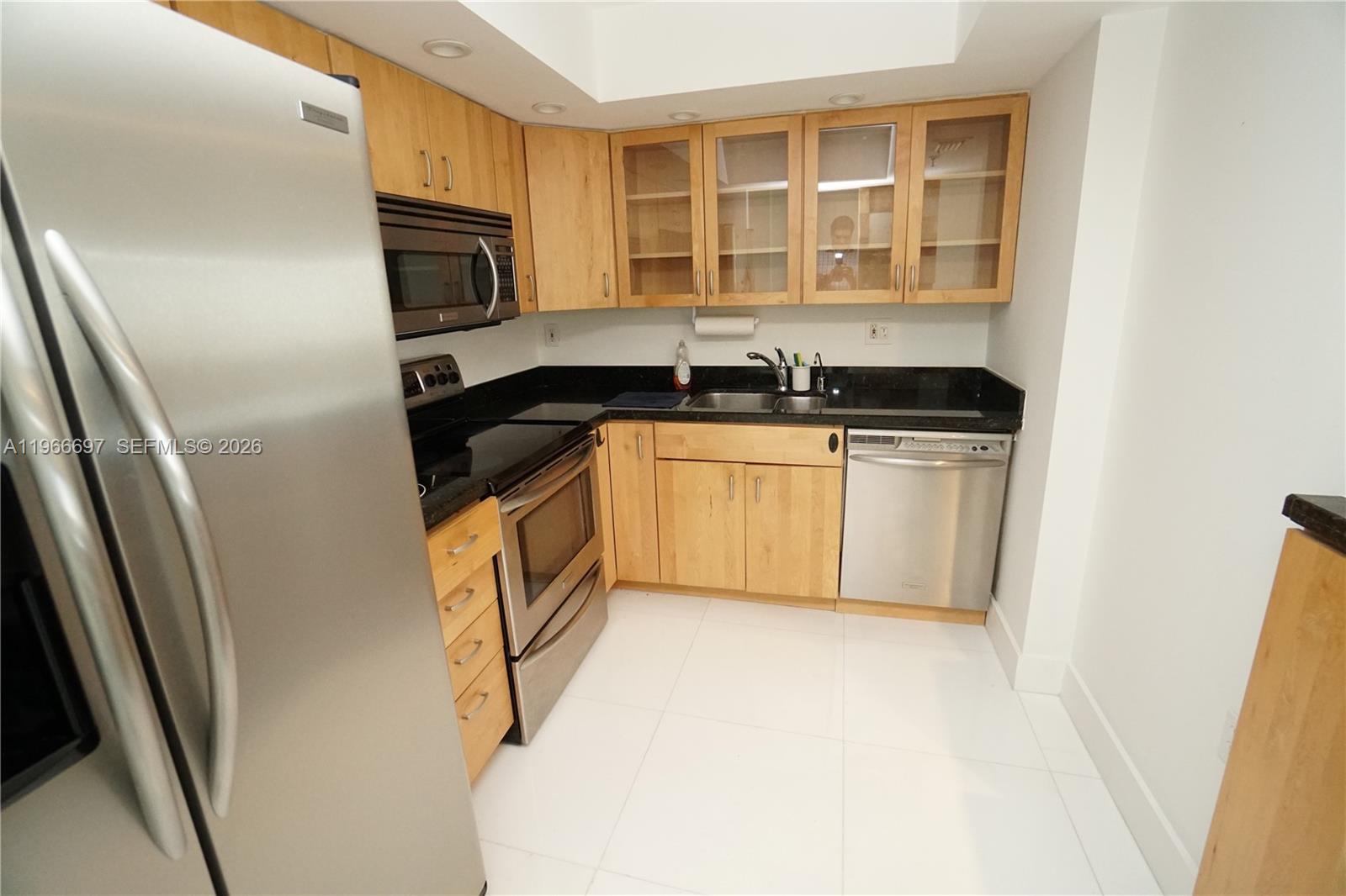 A kitchen features light wood cabinets, stainless steel appliances, and black countertops on white tile — 1775 Washington Ave # 11C, Miami Beach FL 33139, MIAMI BEACH, FL