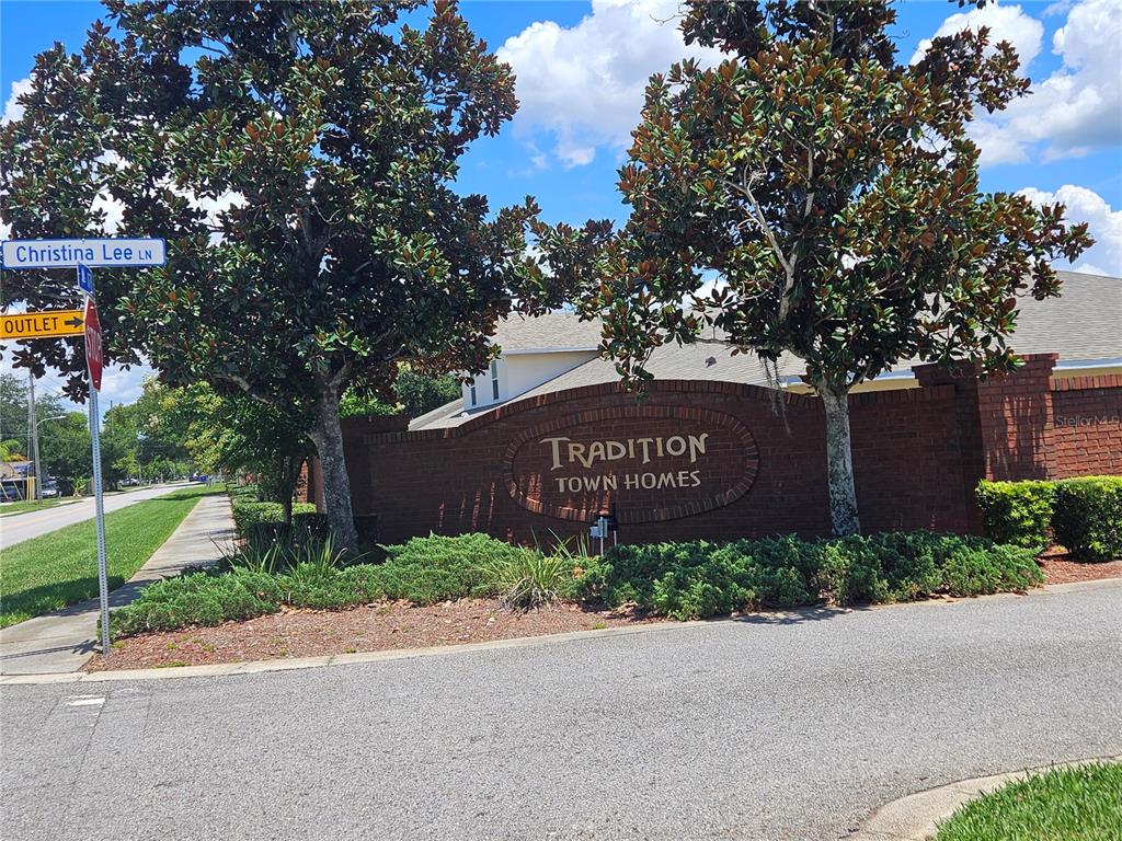 A brick monument sign for Tradition Town Homes stands near a street and large trees — 2696 SHELBY RUTH PLACE, ST CLOUD, FL