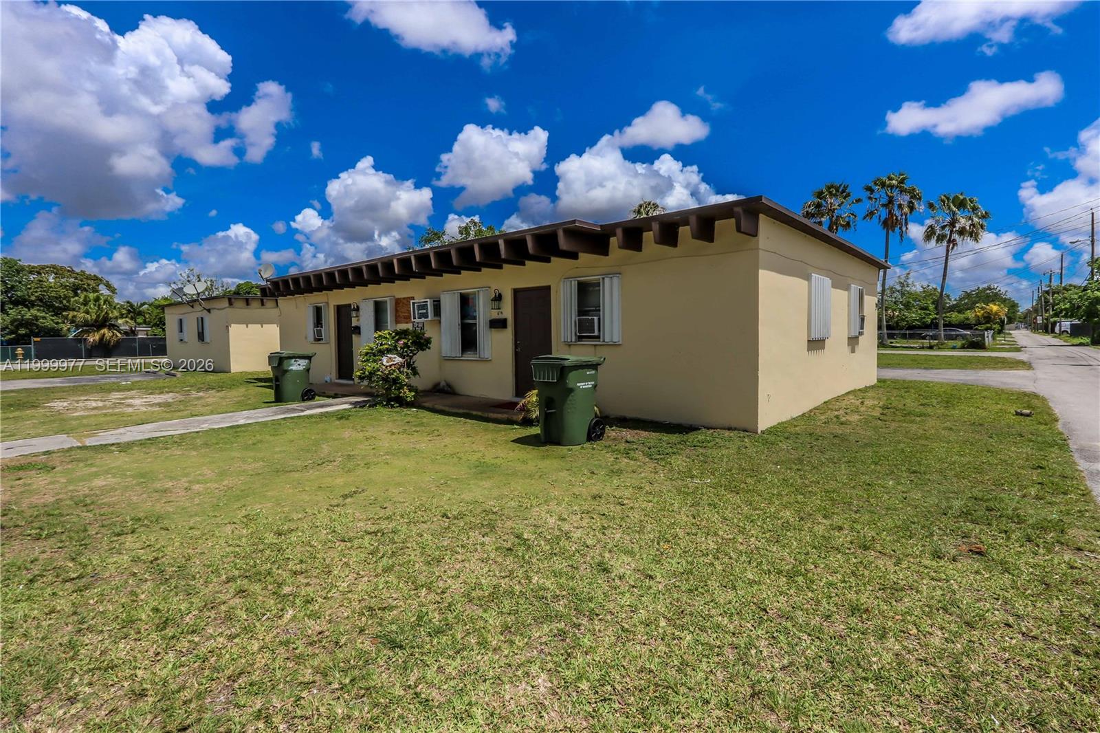 A single-story, light-yellow residential building features a flat roof with exposed rafters and green trash bins — 611 SW 8th Ave, Homestead FL 33030, HOMESTEAD, FL