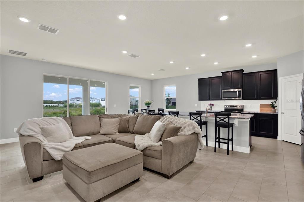 A living room with a sectional sofa and ottoman opens to a kitchen with dark cabinets — 32332 ROSEPINE BOULEVARD, WESLEY CHAPEL, FL