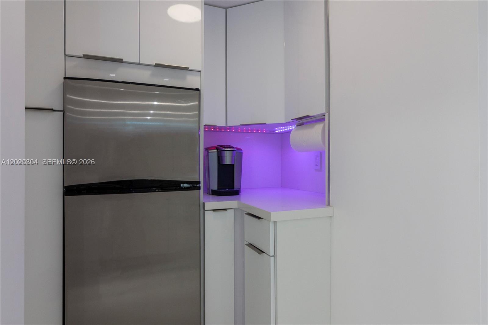 A modern kitchen features white cabinets, a stainless steel refrigerator, and purple under-cabinet lighting — 5445 Collins Ave # 1730, Miami Beach FL 33140, MIAMI BEACH, FL