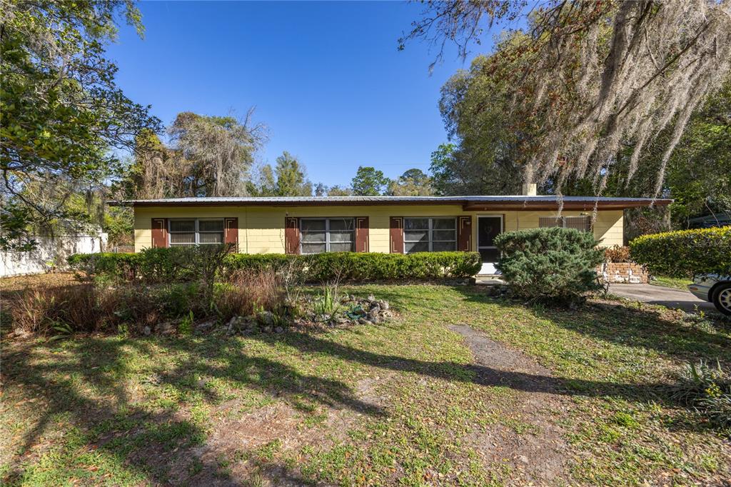 A single-story yellow house with a metal roof and brown shutters sits behind landscaping — 1610 SE 25TH AVENUE, GAINESVILLE, FL