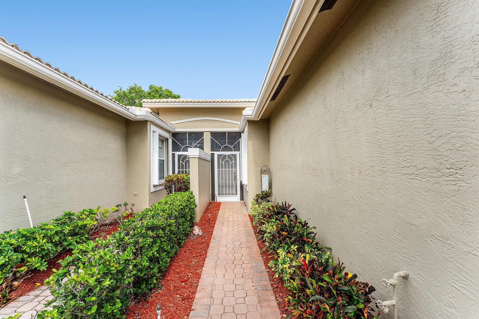 A paved walkway leads to a front entrance framed by stucco walls and landscaped hedges — 10760 Royal Caribbean Circle, Boynton Beach FL 33437, BOYNTON BEACH, FL