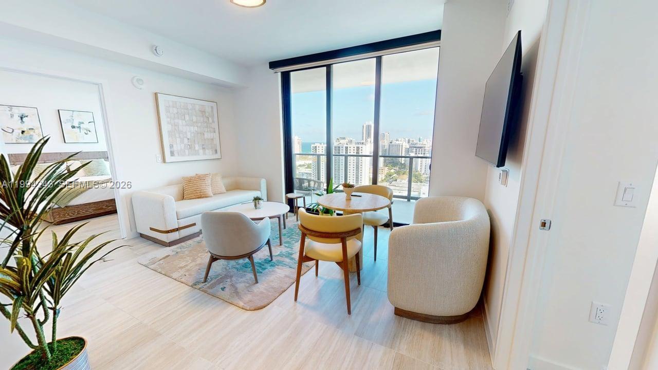 A modern living room features a sofa, dining area, and floor-to-ceiling windows overlooking a city — 580 72nd St # 1708, Miami Beach FL 33141, MIAMI BEACH, FL
