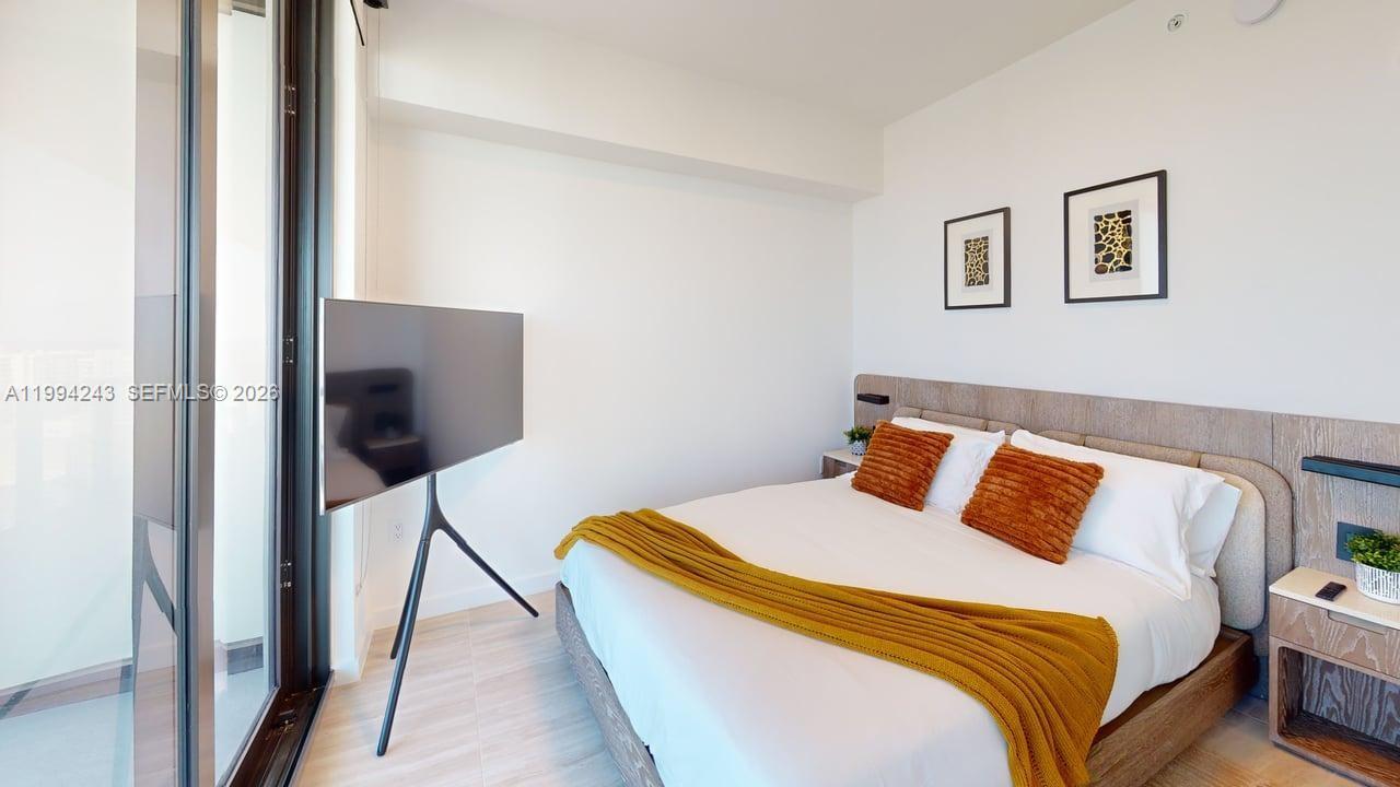 A modern bedroom features a bed with orange pillows, a tripod television, and balcony access — 580 72nd St # 1708, Miami Beach FL 33141, MIAMI BEACH, FL
