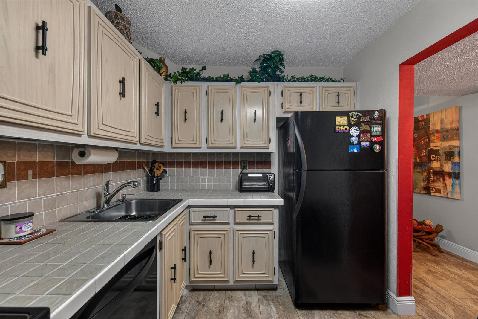 A kitchen features light-colored cabinets, tiled countertops, a black refrigerator, and a red-trimmed doorway — 3671 Environ Boulevard # 169, Lauderhill FL 33319, LAUDERHILL, FL
