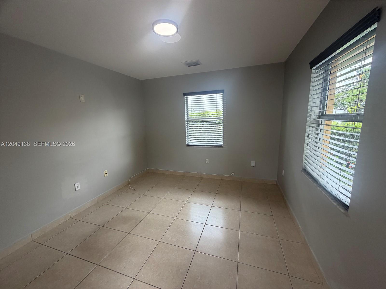 An empty room features light gray walls, beige tile flooring, and two windows with blinds — PALMETTO BAY, FL